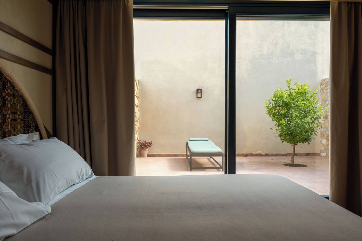 Un dormitorio con cama, una mesita, una planta en maceta y vistas a un patio.