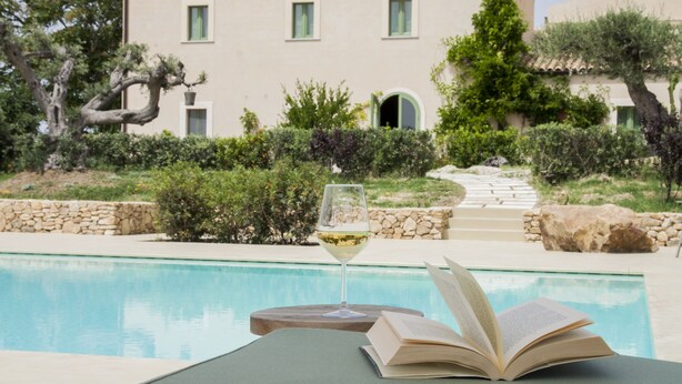 A poolside setting with a book and a glass of white wine on a table, a stone wall, and a building with a tiled roof in the background. - Fontes Episcopi (Aragona)
