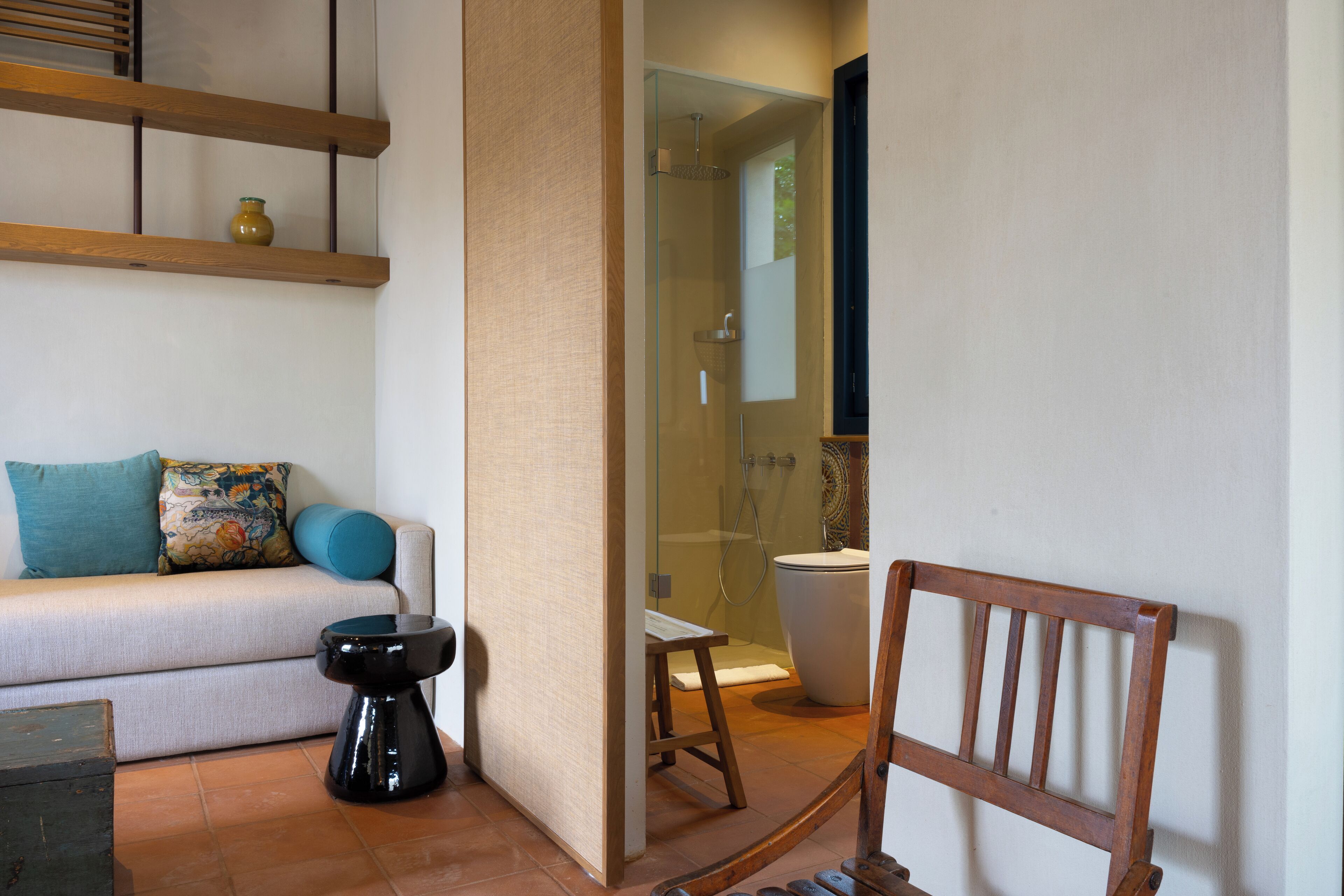 Una stanza con un divano, uno sgabello, una sedia di legno, un box doccia in vetro e un water.