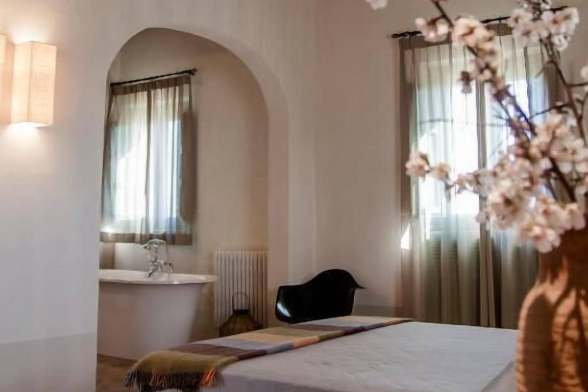Un dormitorio con una cama, una silla, un florero con flores, una ventana con cortinas y un techo de madera.