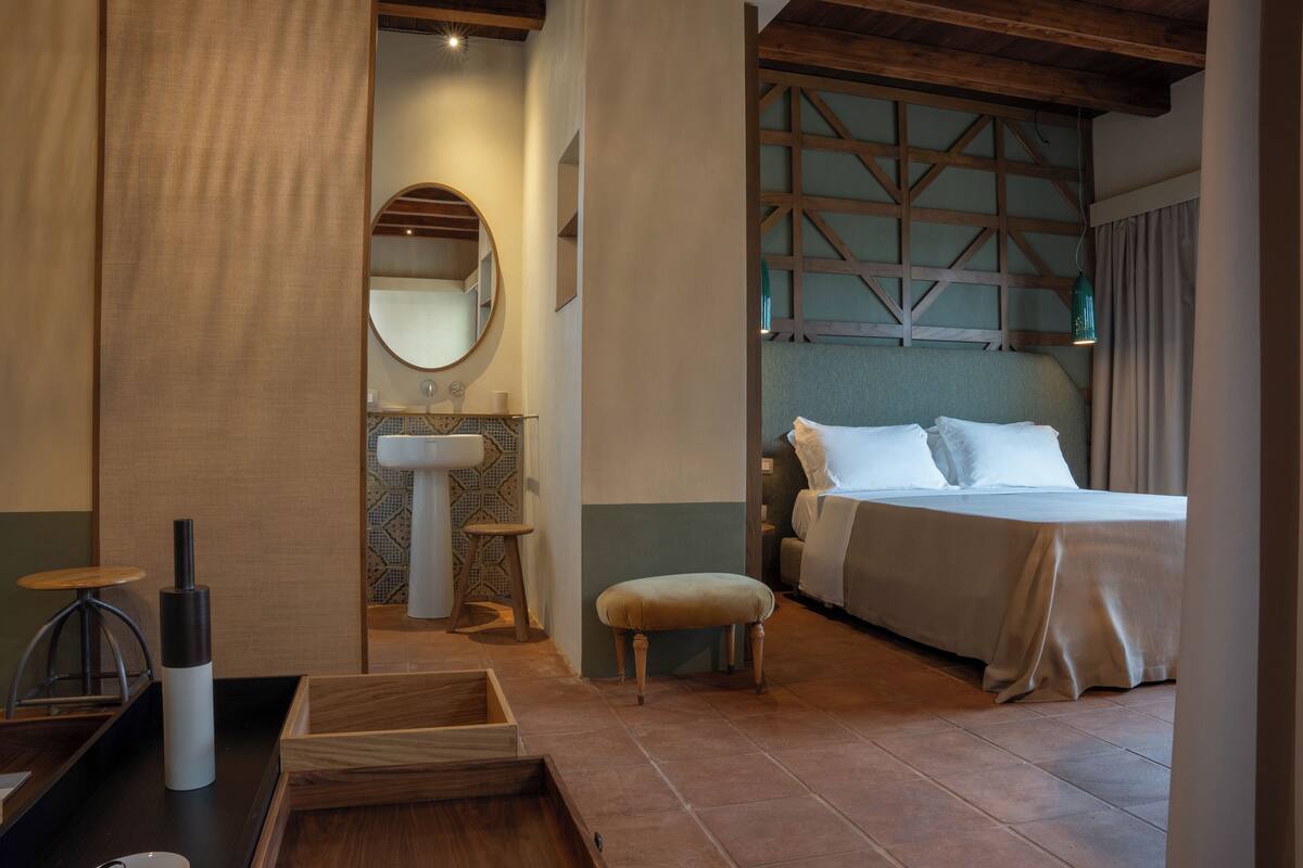 Un dormitorio con cama, una mesita de noche, un espejo, un taburete de madera, una pequeña mesa con una botella y una estantería fijada a la pared.