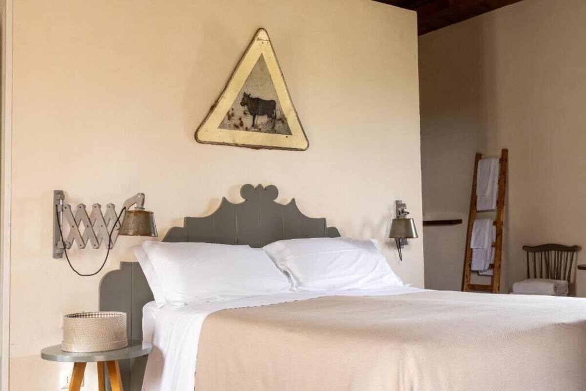 Un dormitorio con una cama, un cuadro triangular en la pared, lámparas de noche, una pequeña mesa redonda con una cesta y una escalera de madera apoyada contra la pared.