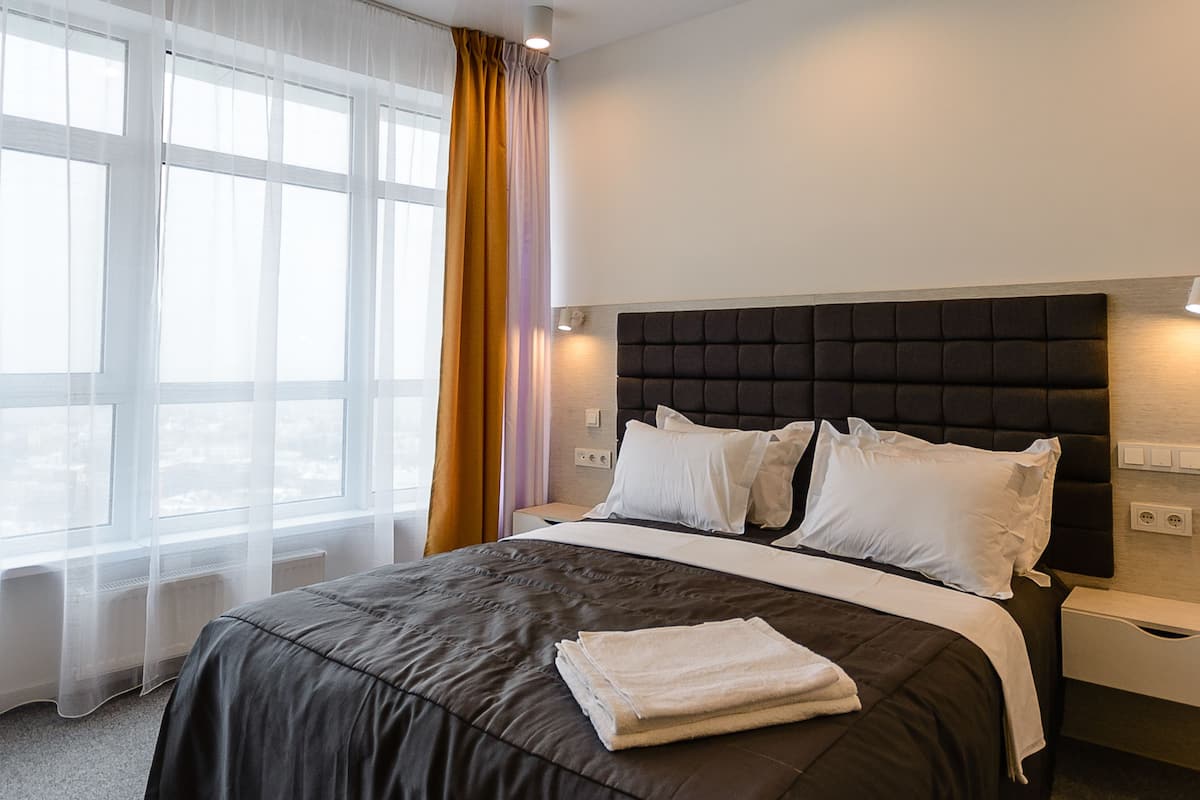 Habitación de hotel con una cama grande, una ventana con cortinas translúcidas, una mesita de noche y una lámpara fijada a la pared.