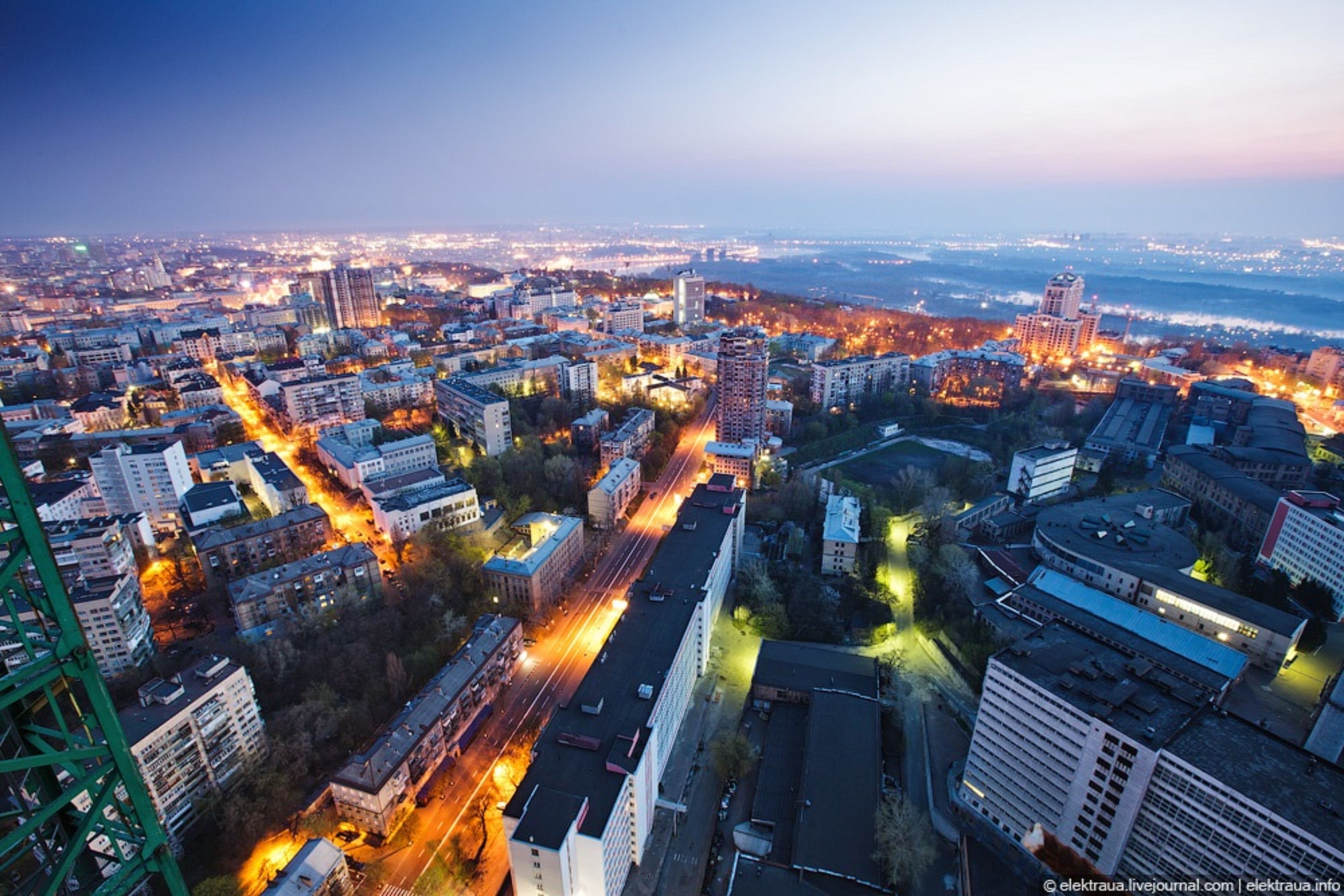A cityscape at dusk with illuminated buildings and a river in the background.