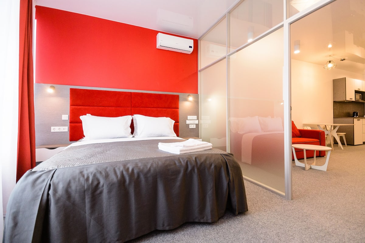 Una habitación de hotel moderna con una pared de acento roja, una cama grande con ropa blanca, un separador de vidrio y una zona de kitchenette.