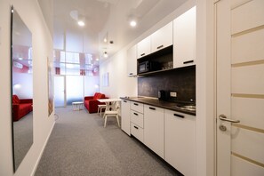 Cozinha moderna com armários brancos, micro-ondas e pia de aço inoxidável. Há uma pequena área de refeições com mesa e cadeiras, e um sofá vermelho ao fundo.