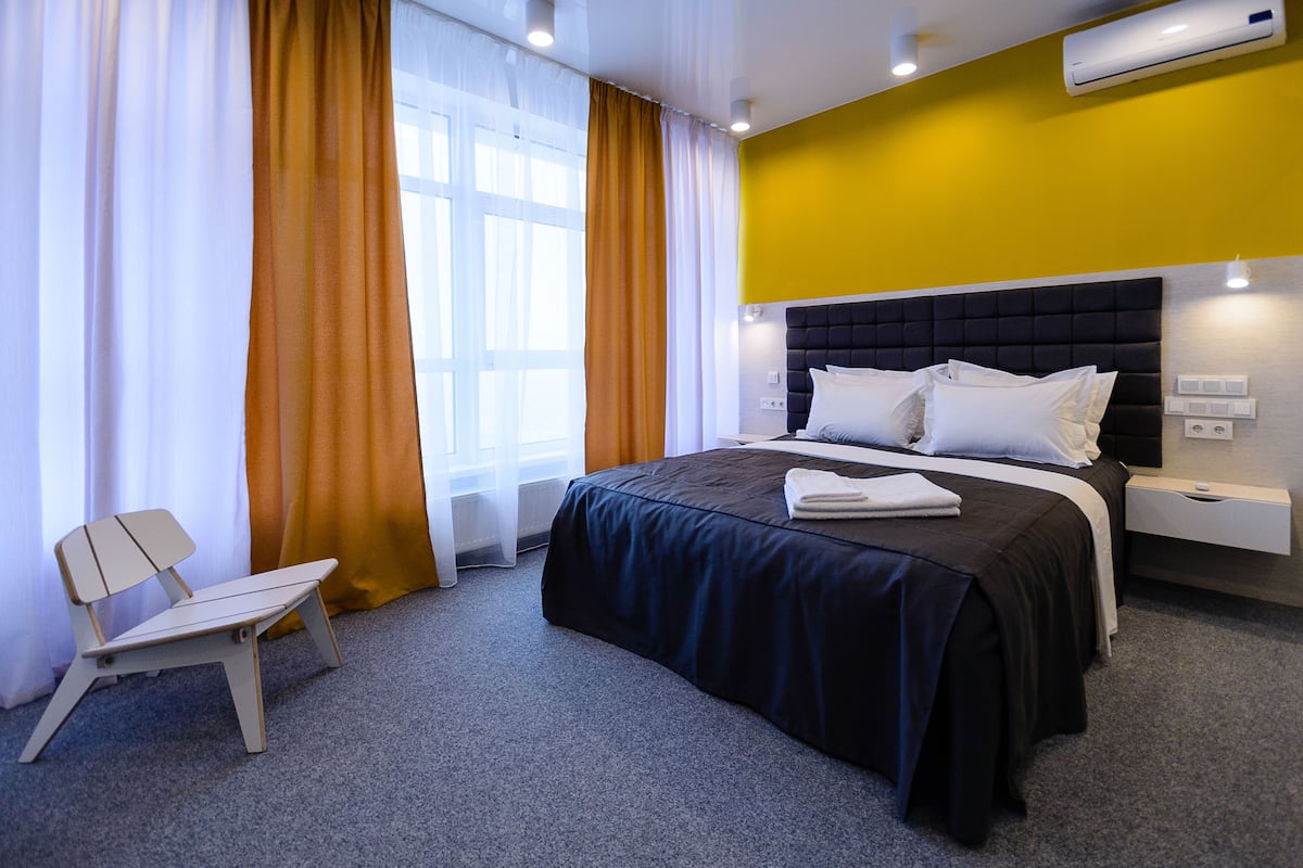 Una habitación de hotel moderna con una cama grande, una mesita de noche, una silla y una ventana con cortinas.