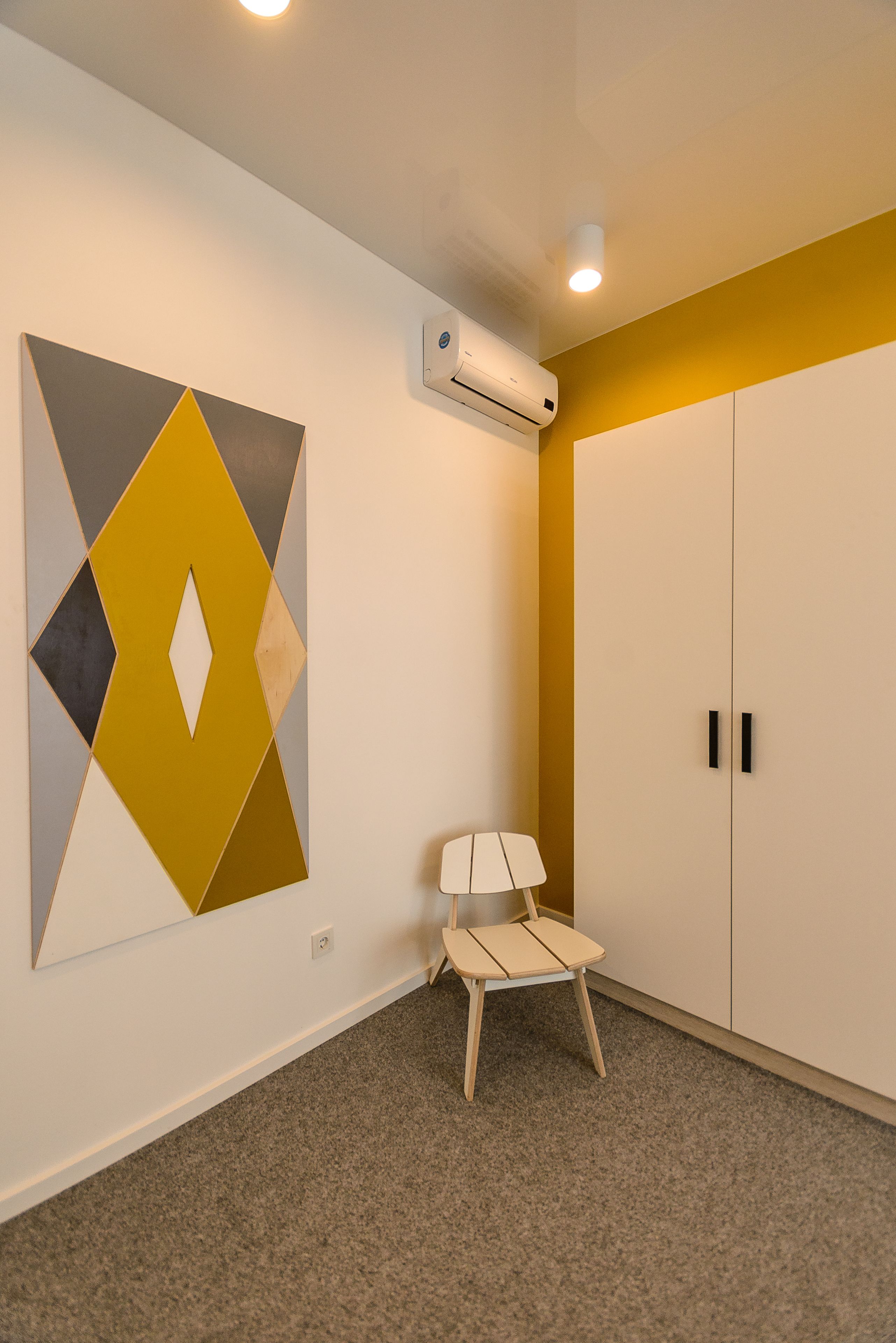 A room with a geometric wall art, a white wardrobe, a small wooden chair, and an air conditioning unit.