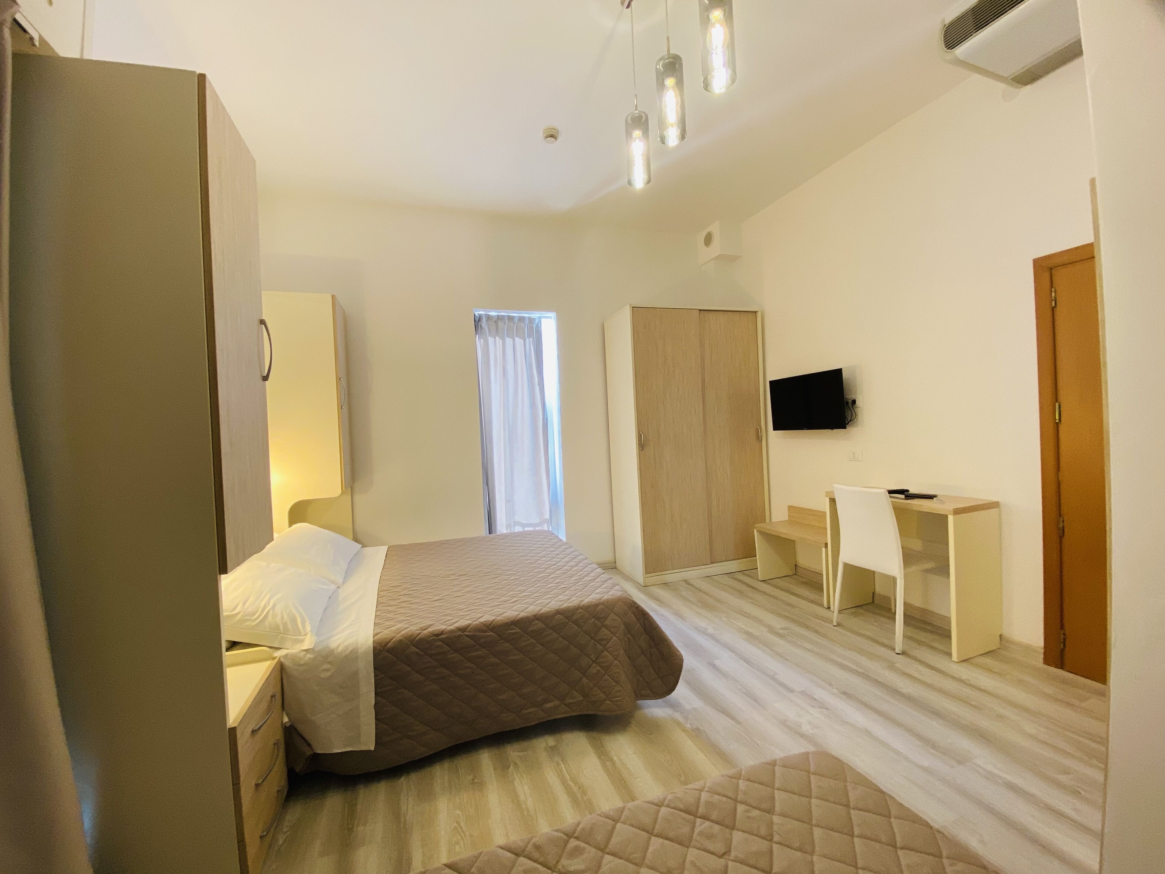 Ein Schlafzimmer mit Bett, Schreibtisch, Stuhl, Kleiderschrank, Fernseher und einem Fenster mit Vorhängen.