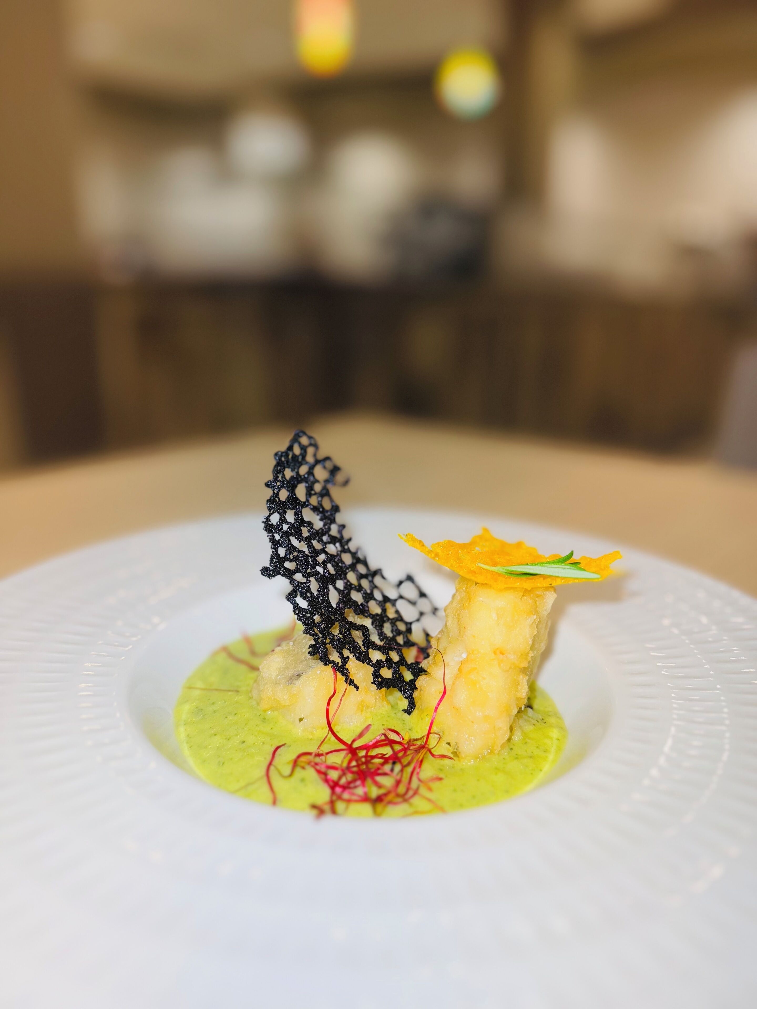 A plate with a food presentation including a piece of food with a yellow crust, a black net-like structure, and green sauce with red strands.