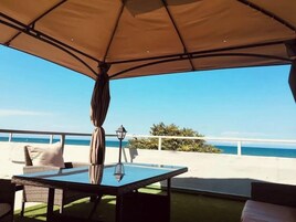 Un área de estar al aire libre con una mesa y sillas de vidrio, un paraguas color beige y vistas al mar.