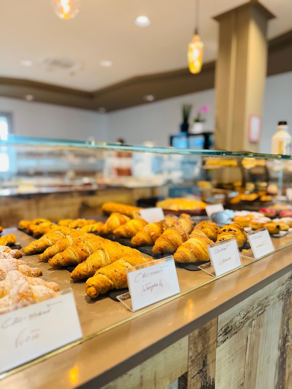 Mostrador de panadería con diversos tipos de pasteles, incluyendo cruasanes y otras opciones horneadas, con etiquetas que indican sus nombres.