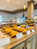Mostrador de panadería con diversos tipos de pasteles, incluyendo cruasanes y otras opciones horneadas, con etiquetas que indican sus nombres.