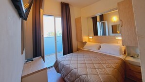 Habitación de hotel con cama, escritorio, televisor, espejo y puerta corrediza a un balcón.