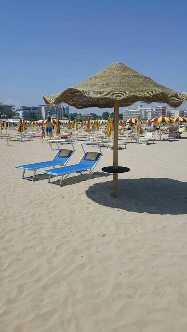 Una playa con tumbonas y sombrillas, una estructura de sombrilla de paja y edificios al fondo.