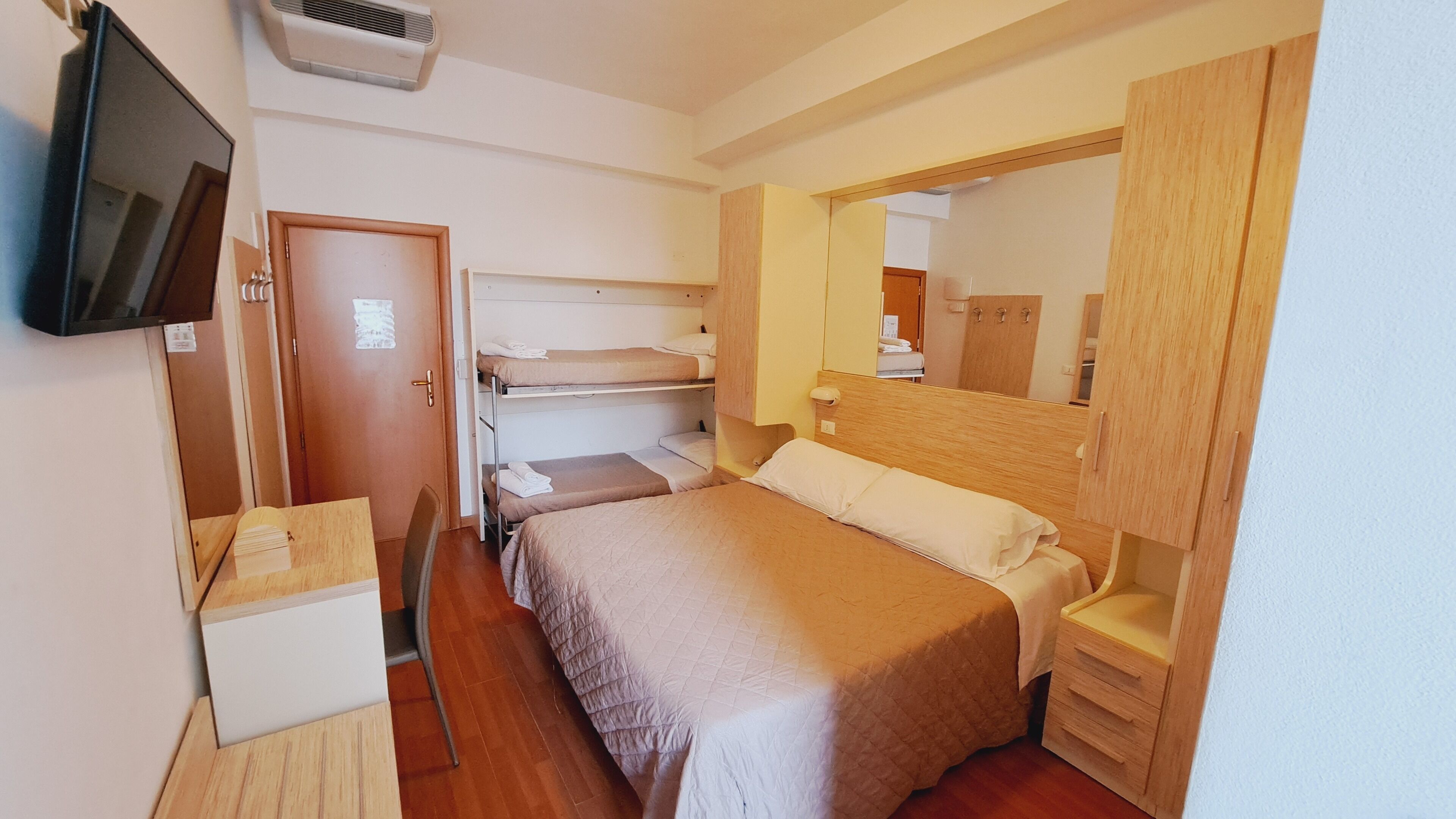 A compact hotel room with bunk beds, a wooden wardrobe, a small desk, and a television.