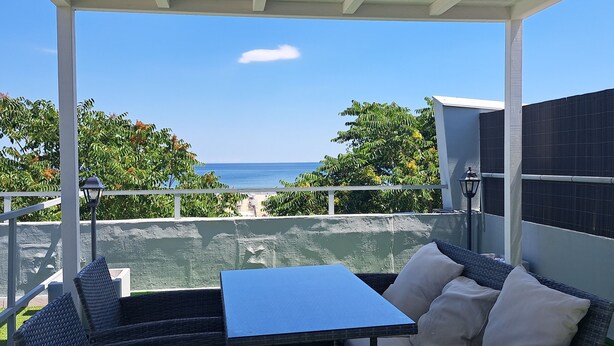 Un patio techado con muebles de mimbre y una mesa, con vistas a un cuerpo de agua y follaje.