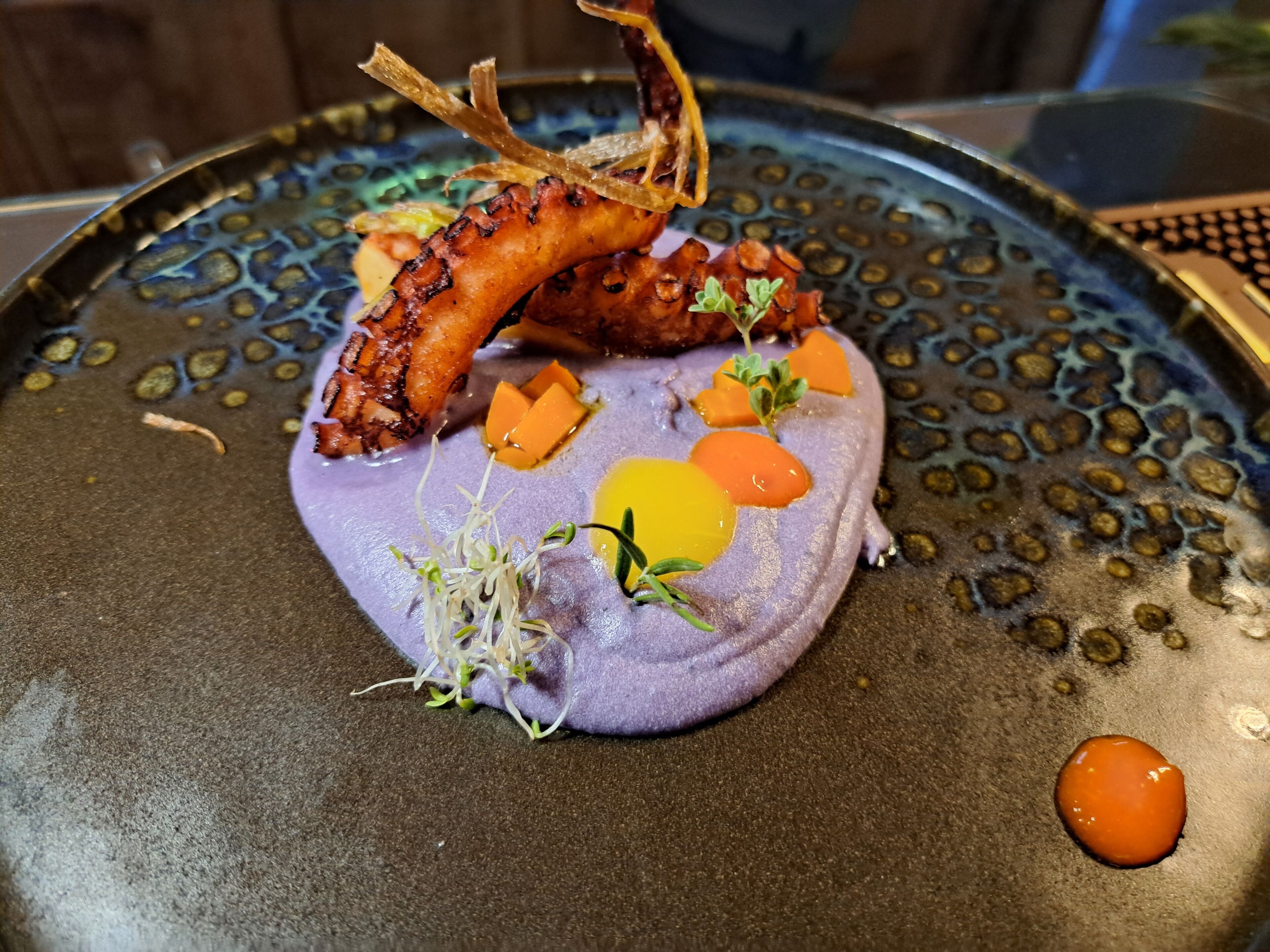 A plate with a purple puree, orange cubes, sprouts, and a grilled octopus.