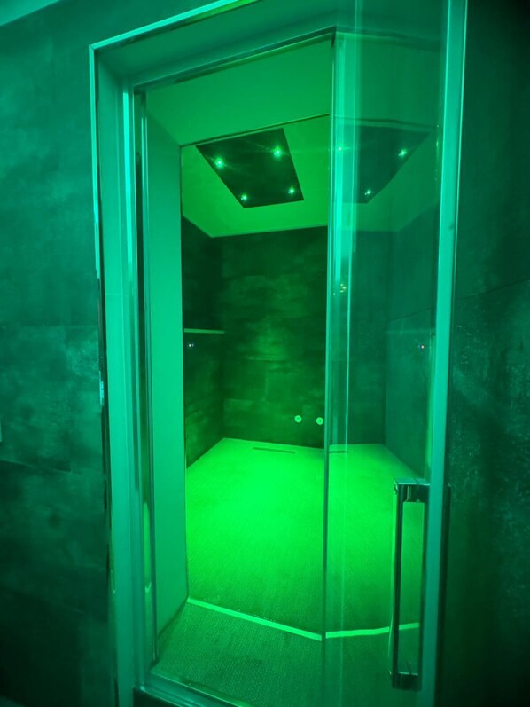 Una habitación con paredes de vidrio, techo iluminado en verde y un ventanal pequeño.