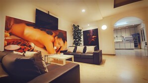 A modern living room with a sofa, a coffee table, and a large poster of a spa scene. There is a TV mounted on the wall, a plant, and a kitchen area in the background. - Hotel Butterfly & YoUnique Spa (Rimini)