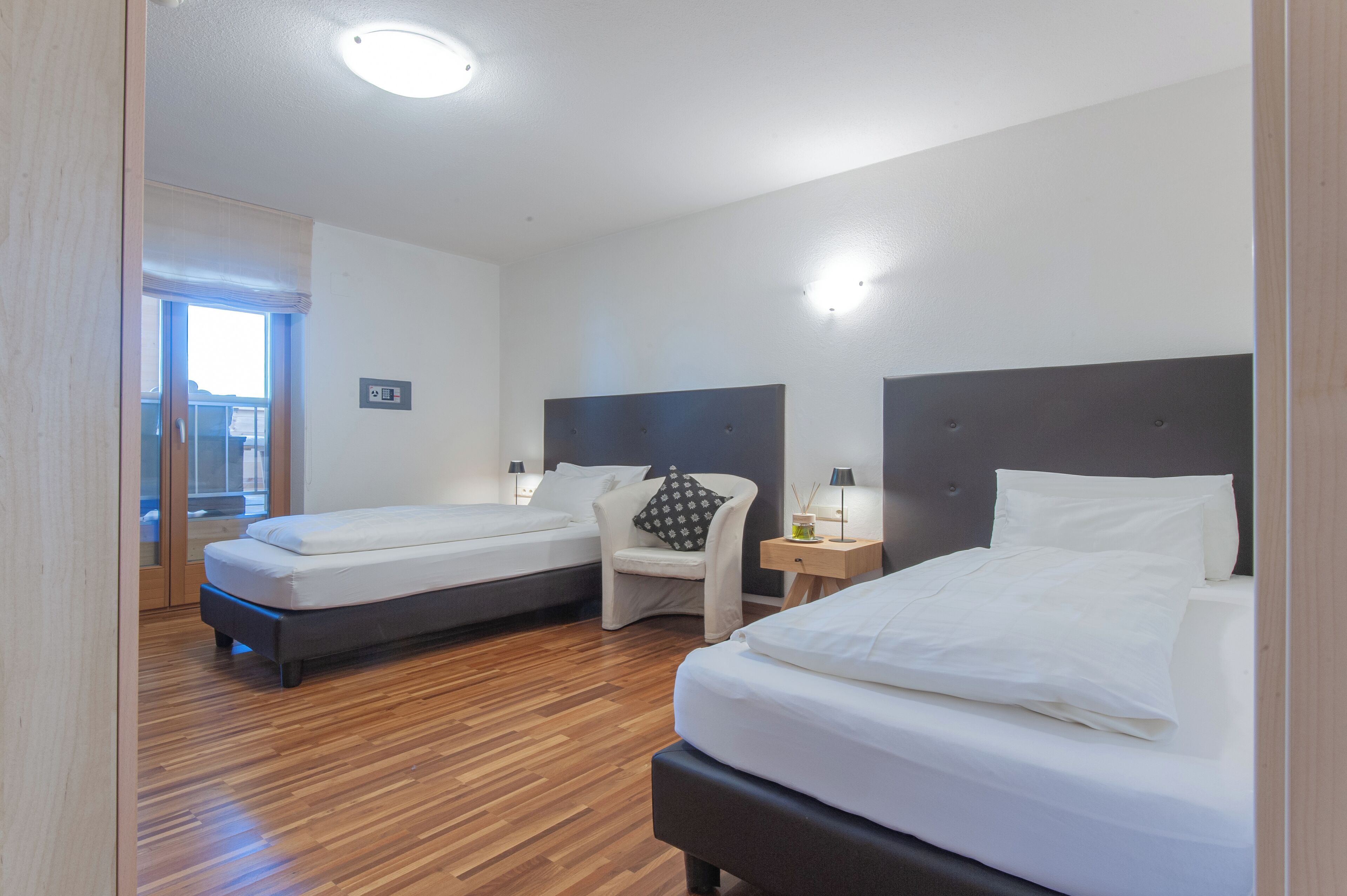 A hotel room with two single beds, a wooden floor, a chair, a nightstand with a lamp, and a window with a view.