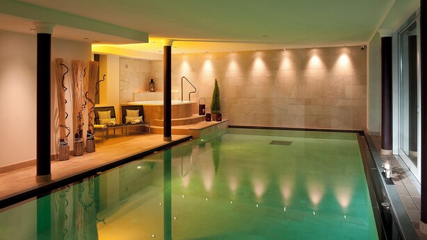 A spacious indoor swimming pool with a clear water surface, a seating area with a bench and cushions, and a wall with recessed lighting. - Hotel Italia (Corvara in Badia)