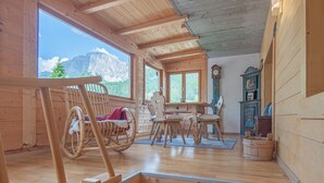 A wooden cabin interior with a dining area, rocking chairs, and a view of mountains through large windows. - Hotel Italia (Corvara in Badia)