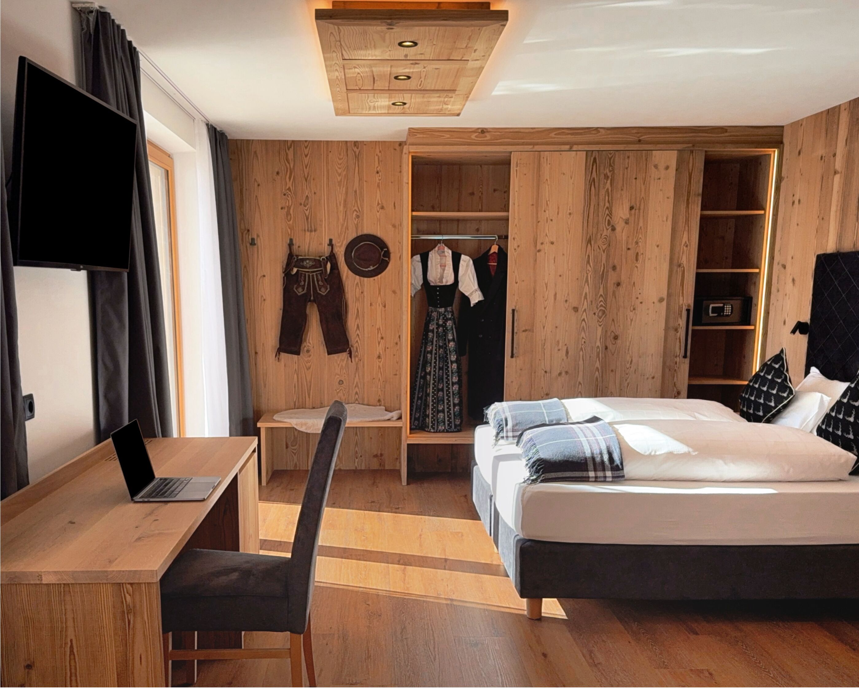 Een slaapkamer met een houten garderobe, een bureau met een laptop, een bed met kussens en een televisie aan de muur.