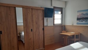 A room with a wooden wardrobe, a desk with a chair, a bed with a white bedspread, and a window with a view of mountains. - Residence Biancaneve (Aprica)