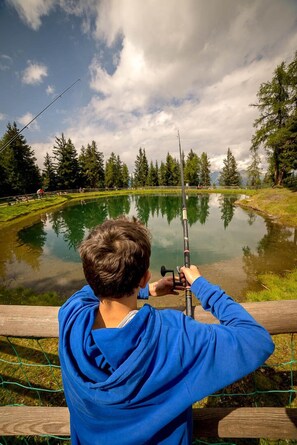 A person fishing by a calm lake surrounded by trees. - Residence Biancaneve (Aprica)
