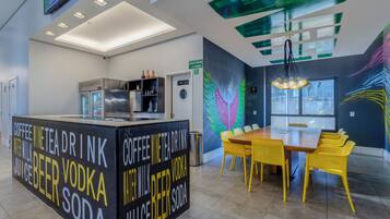 A modern kitchen with a coffee station, a dining area with yellow chairs, and a mural on the wall.