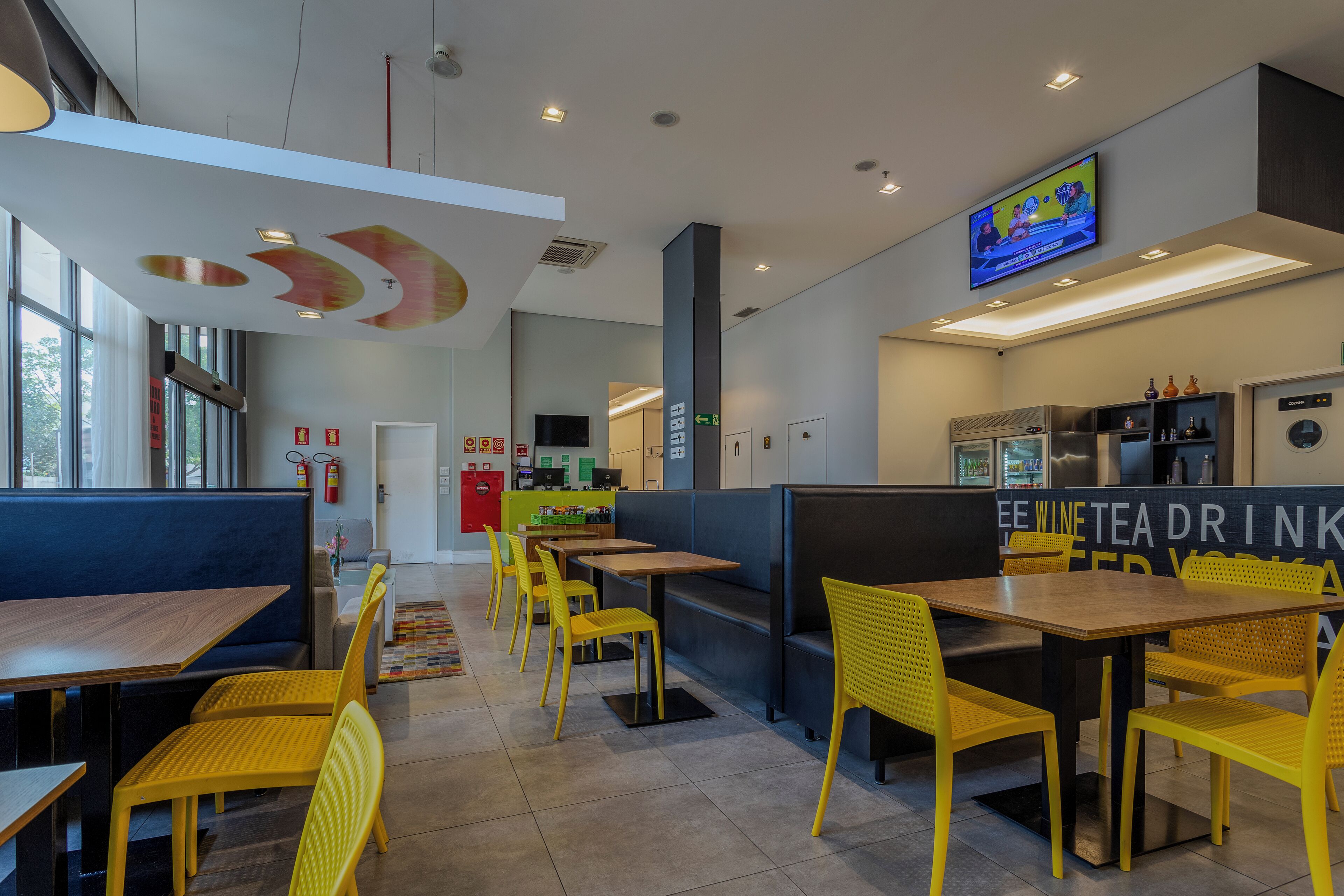 A modern restaurant interior with yellow chairs, wooden tables, and a TV mounted on the wall.