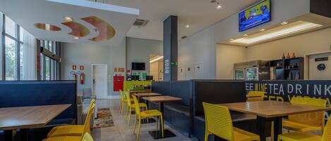 A modern restaurant interior with yellow chairs, wooden tables, and a TV mounted on the wall.