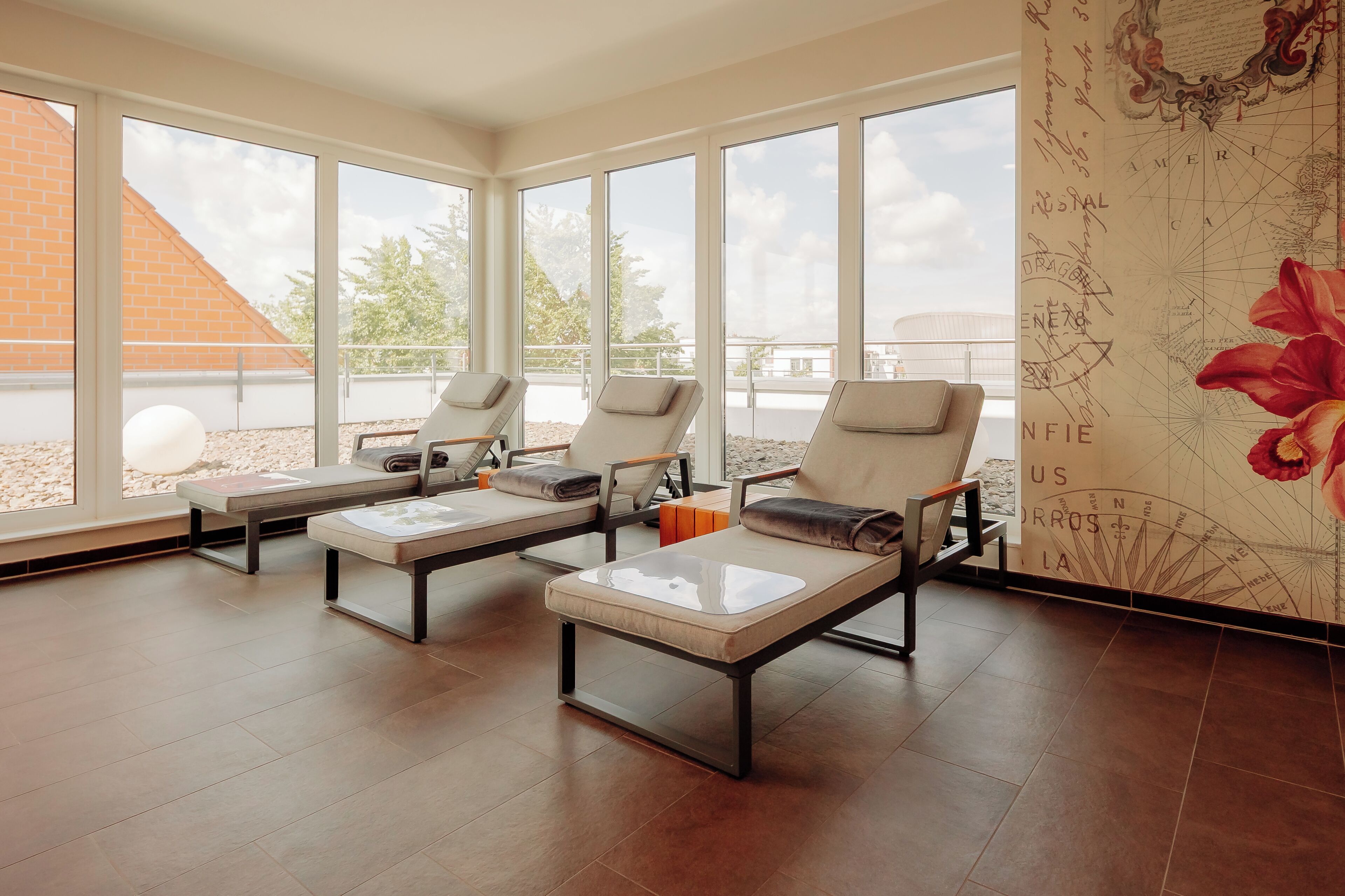 A sunlit room with large windows, three sun loungers, and a wall with a map and decorative elements.