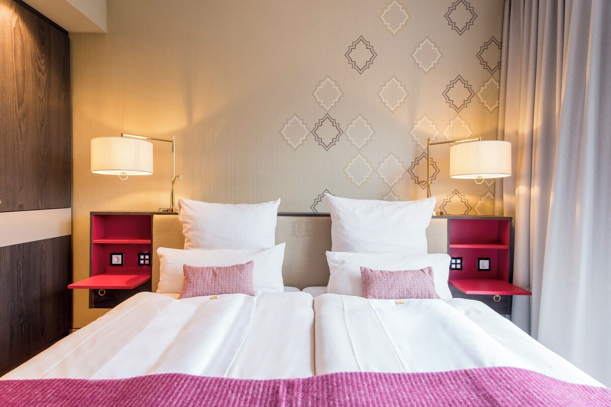 A double bed with white linens, two bedside tables with lamps, and a red shelf unit.