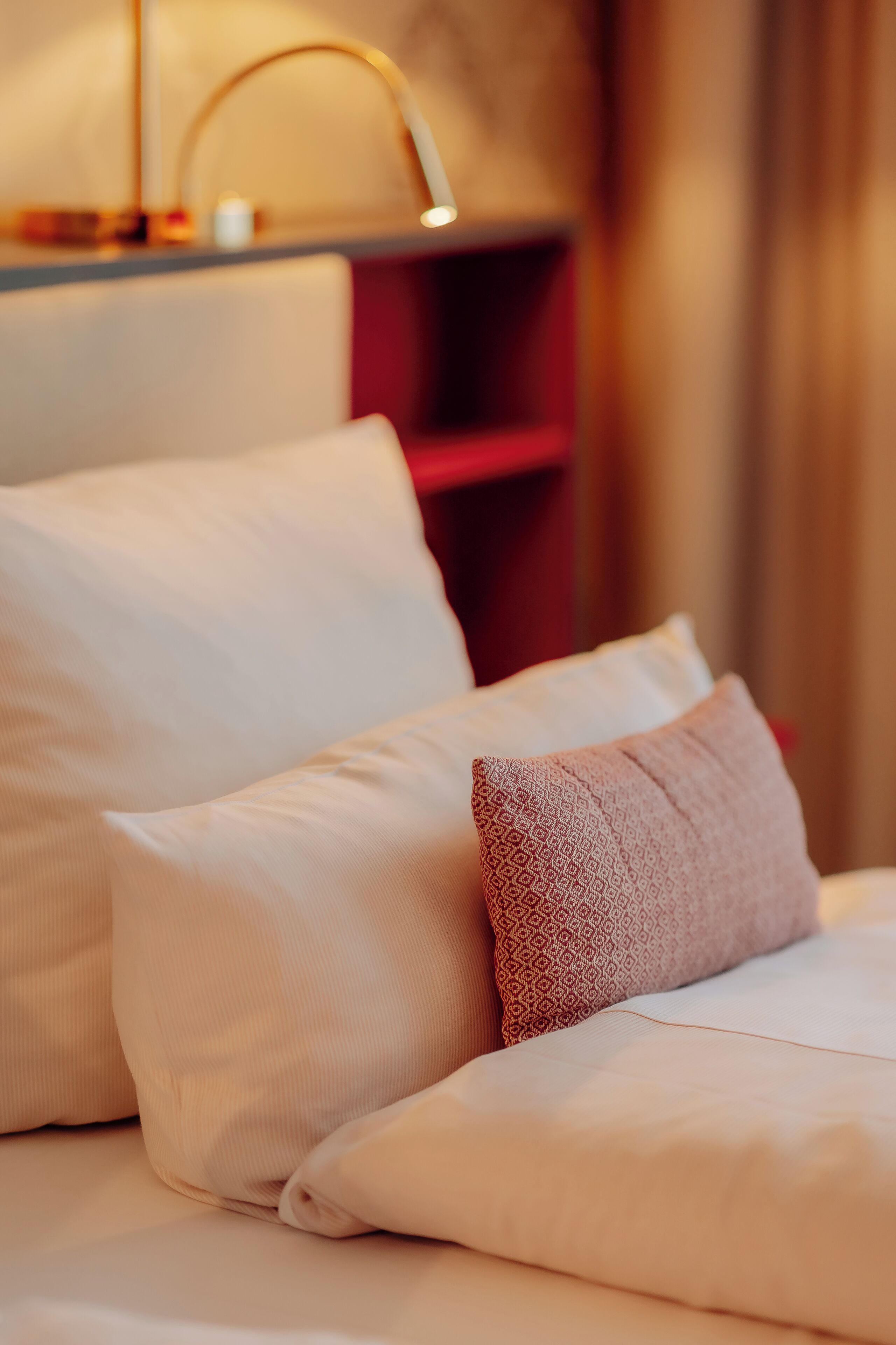 A neatly made bed with white and patterned pillows, a bedside table with a lamp, and a red shelving unit in the background.