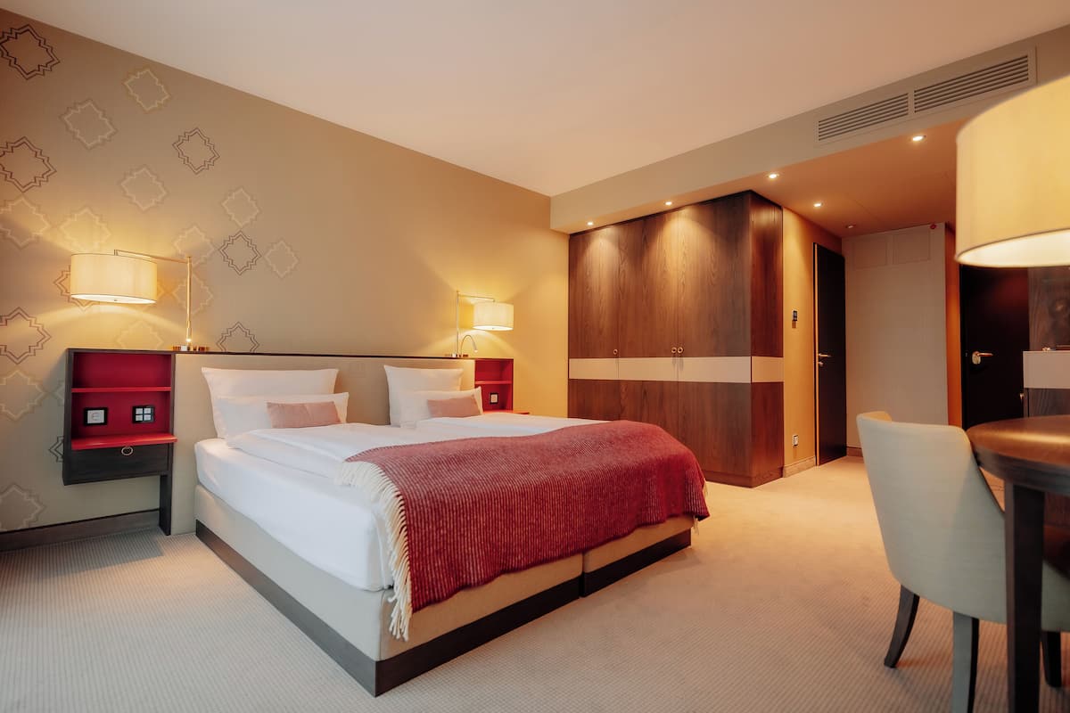 A modern hotel room with a large bed, bedside tables with lamps, a wardrobe, and a desk with a chair.
