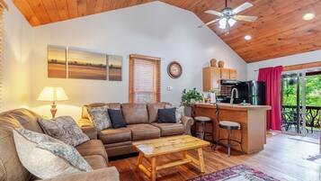 A cozy living area with a wooden ceiling, a sofa, a coffee table, and a kitchen area with a bar and stools.