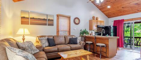 Una sala de estar acogedora con techo de madera, un sofá, una mesa de centro y una zona de cocina con barra y taburetes.