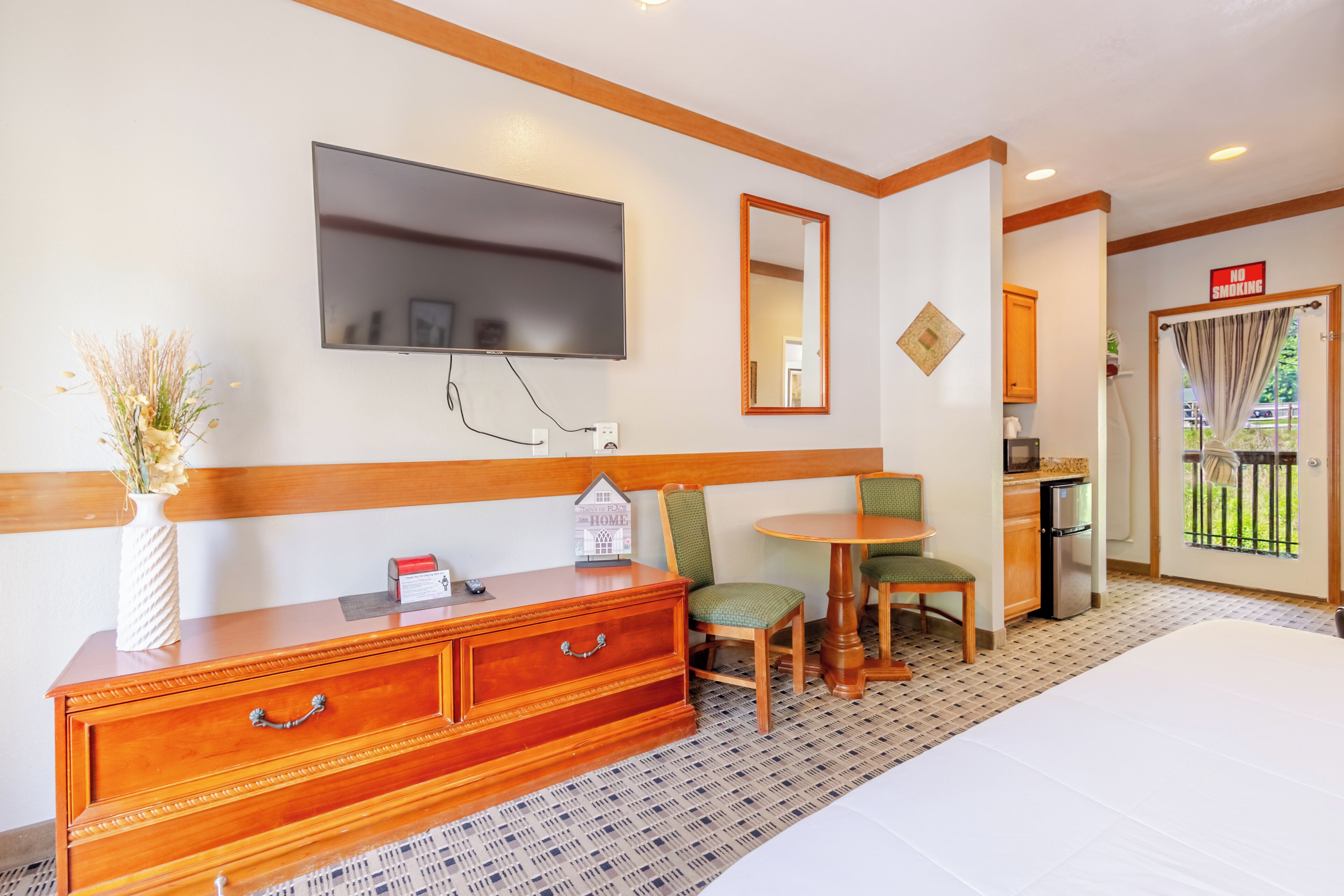 A hotel room with a bed, a wooden dresser, a television, a small table with chairs, a kitchen area, and a balcony with a view.