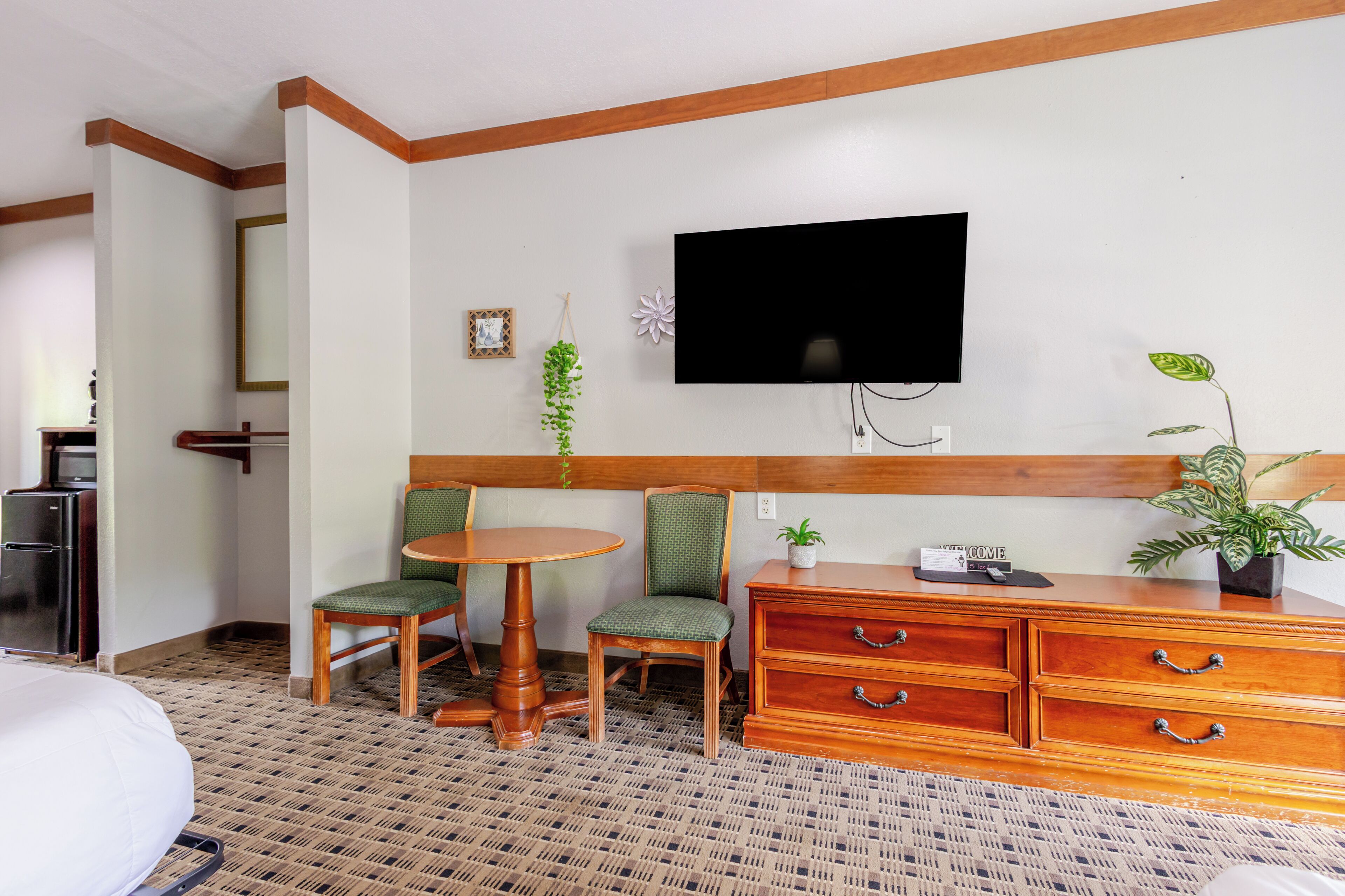 A hotel room with a bed, a desk with a chair, a television mounted on the wall, a dresser, and a potted plant.