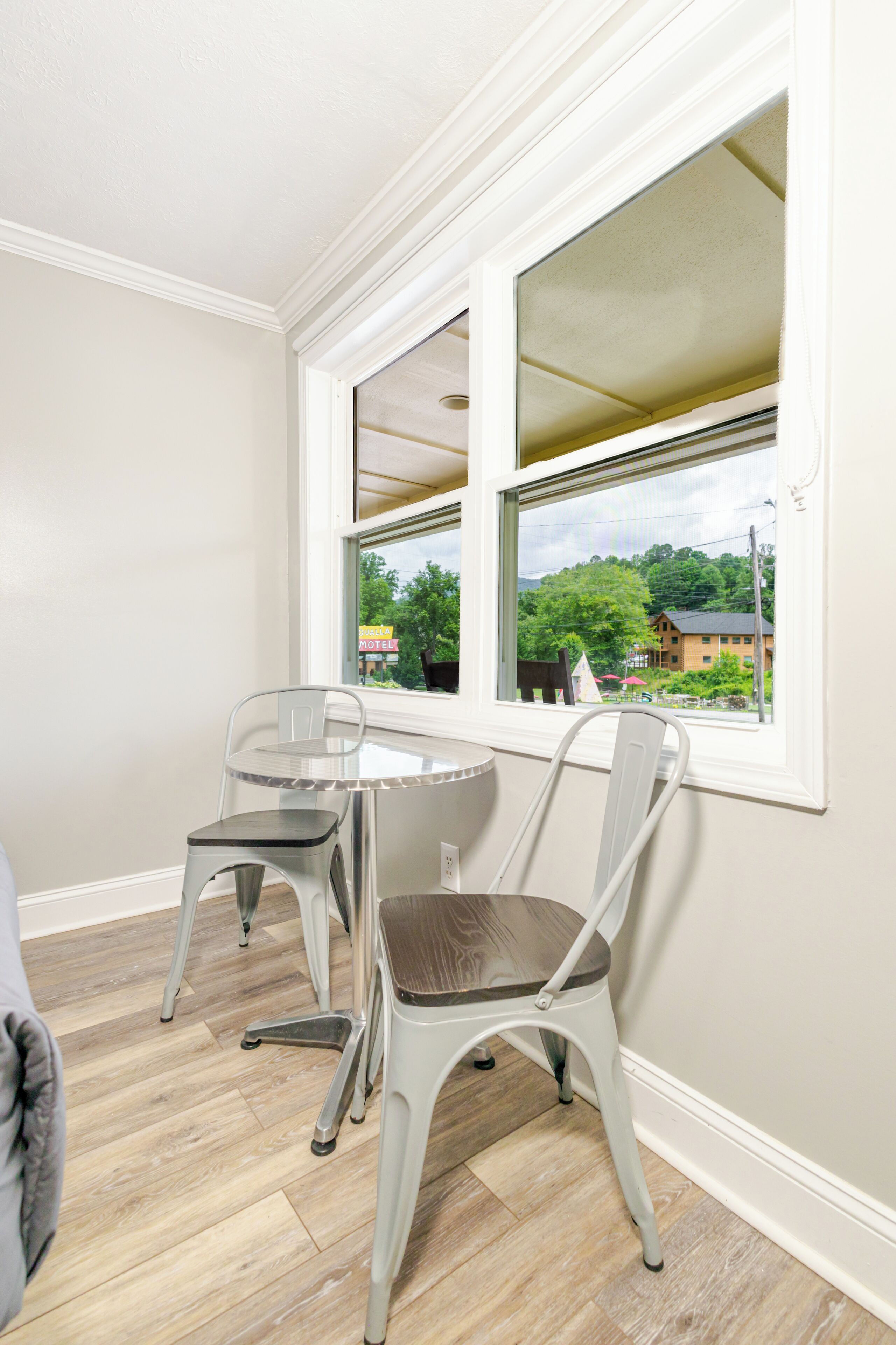 A corner room with a window, a small table, two chairs, and a view of a building and trees outside.