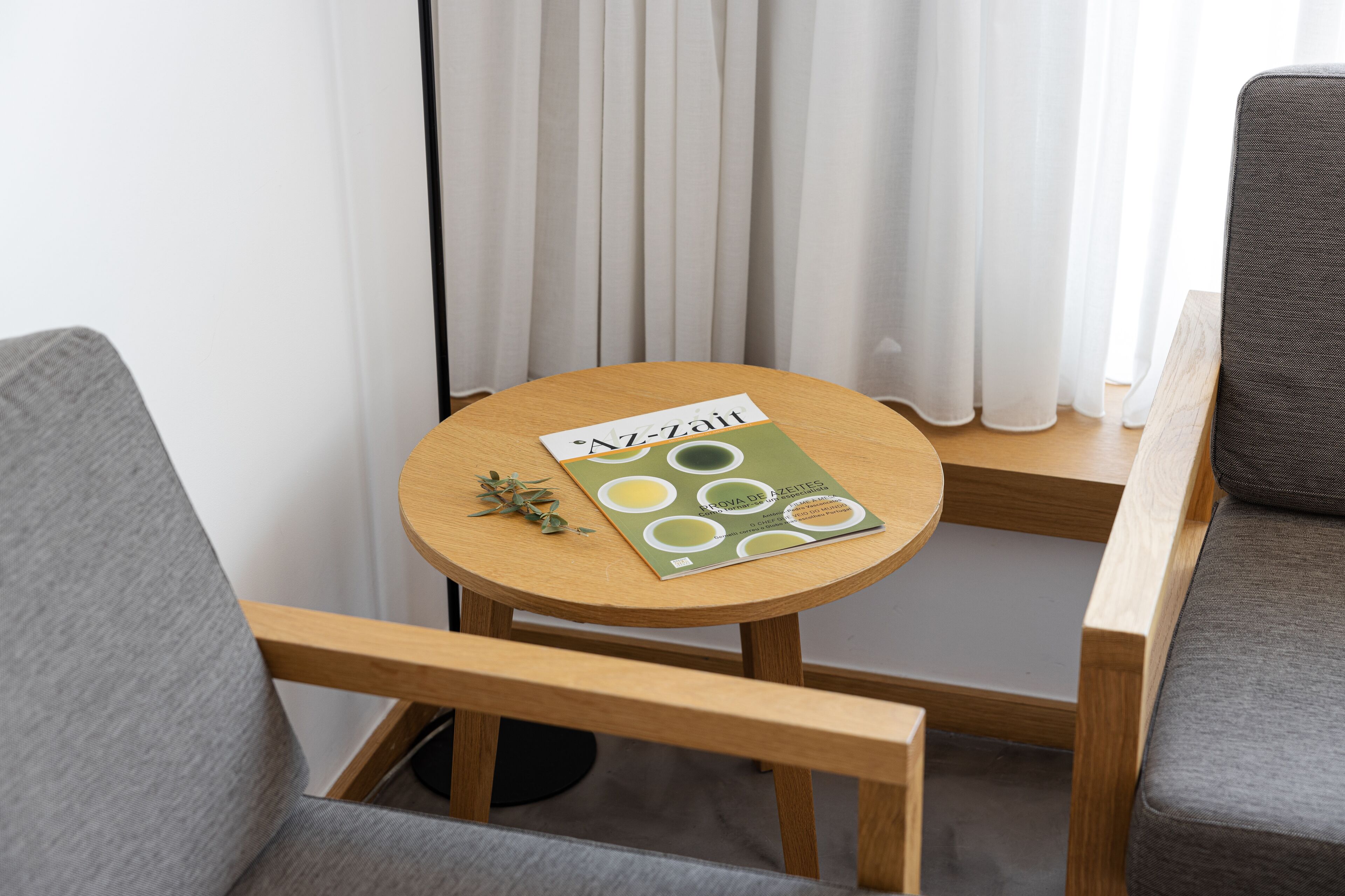 Une petite table ronde en bois, sur laquelle repose un magazine, est placée entre deux fauteuils gris recouverts de tissu.