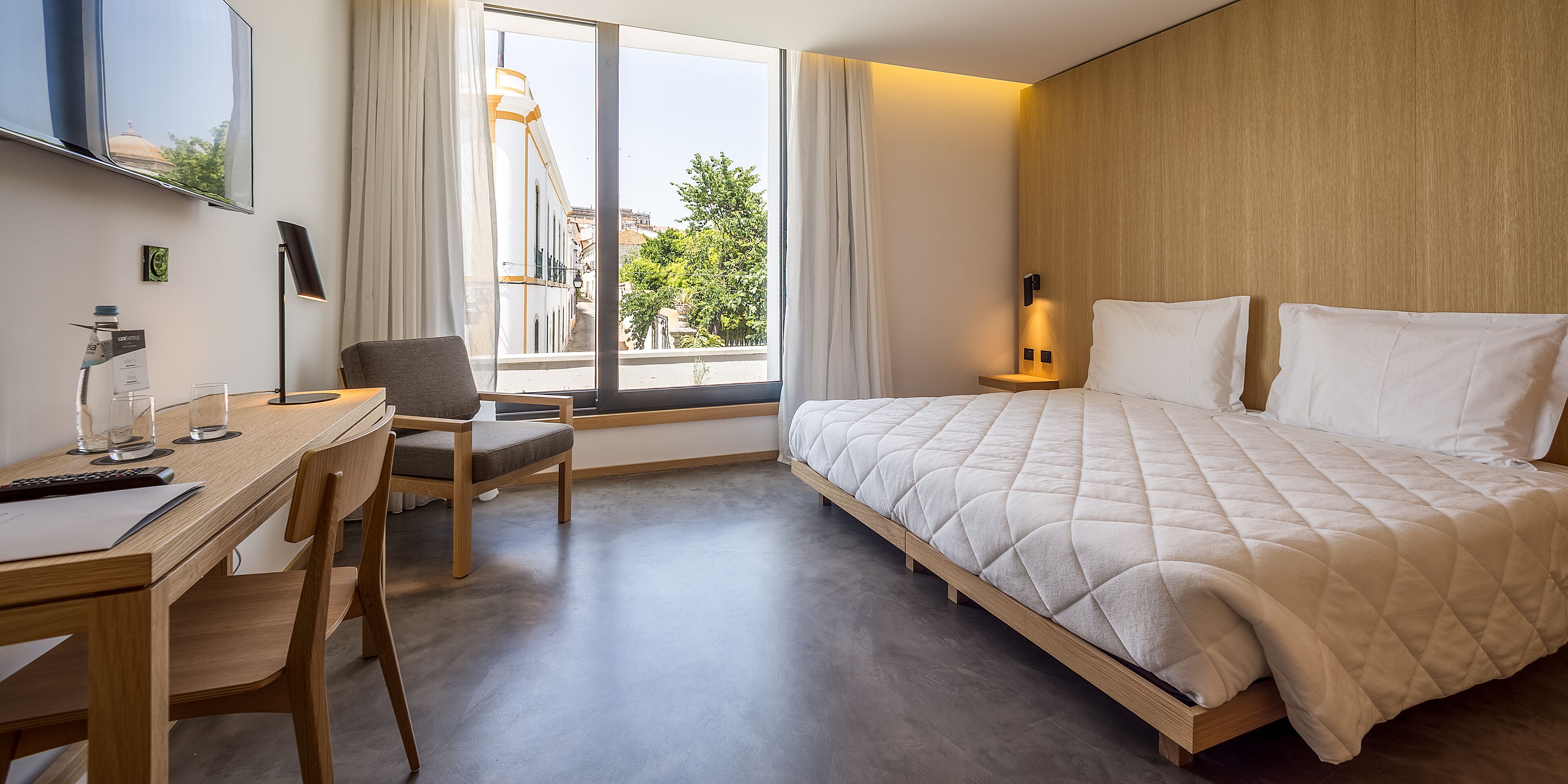 Habitación de hotel con cama, escritorio con silla, televisor montado en la pared y ventana con cortinas.