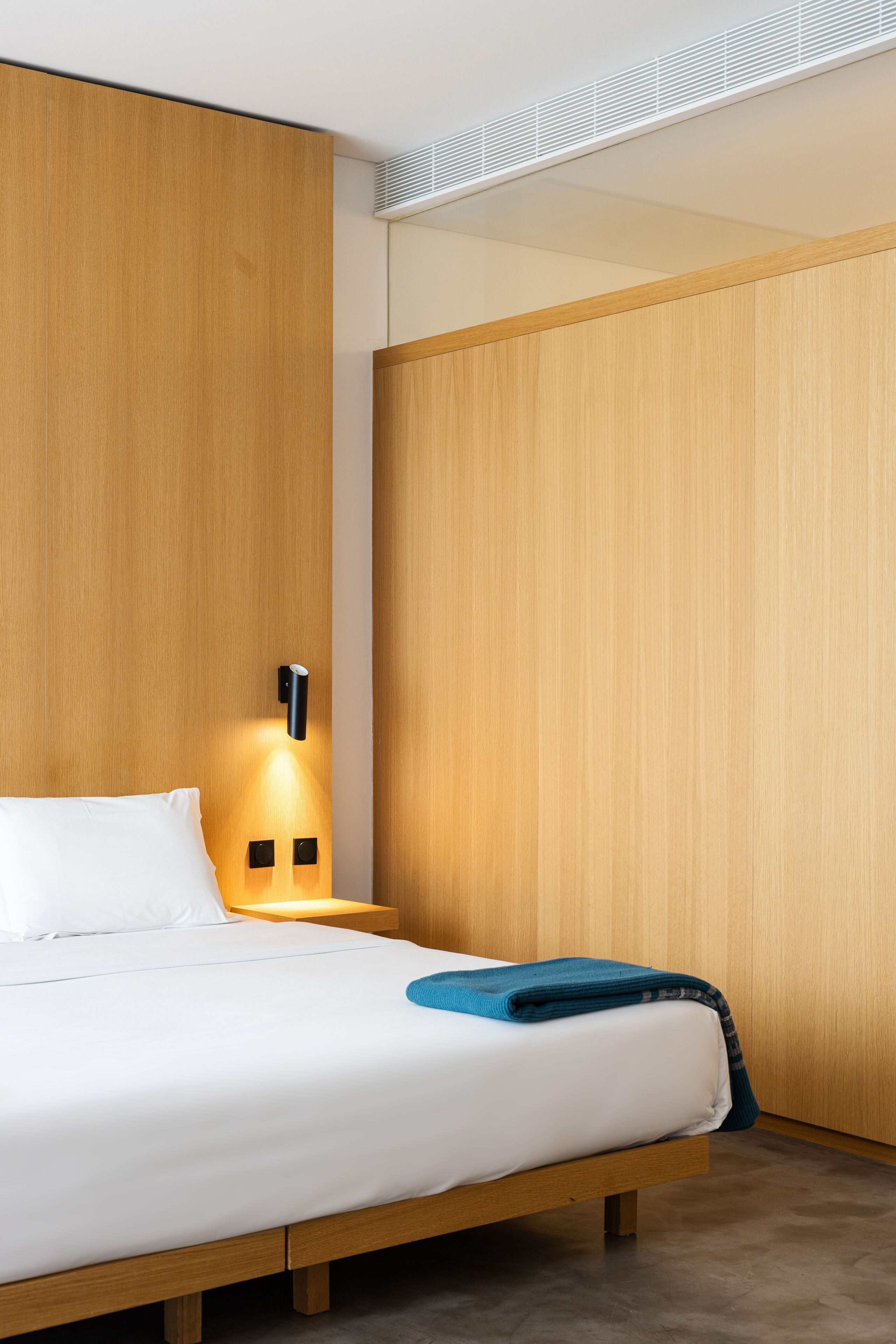 A modern hotel room with a wooden bed, a nightstand with a lamp, and a wardrobe.