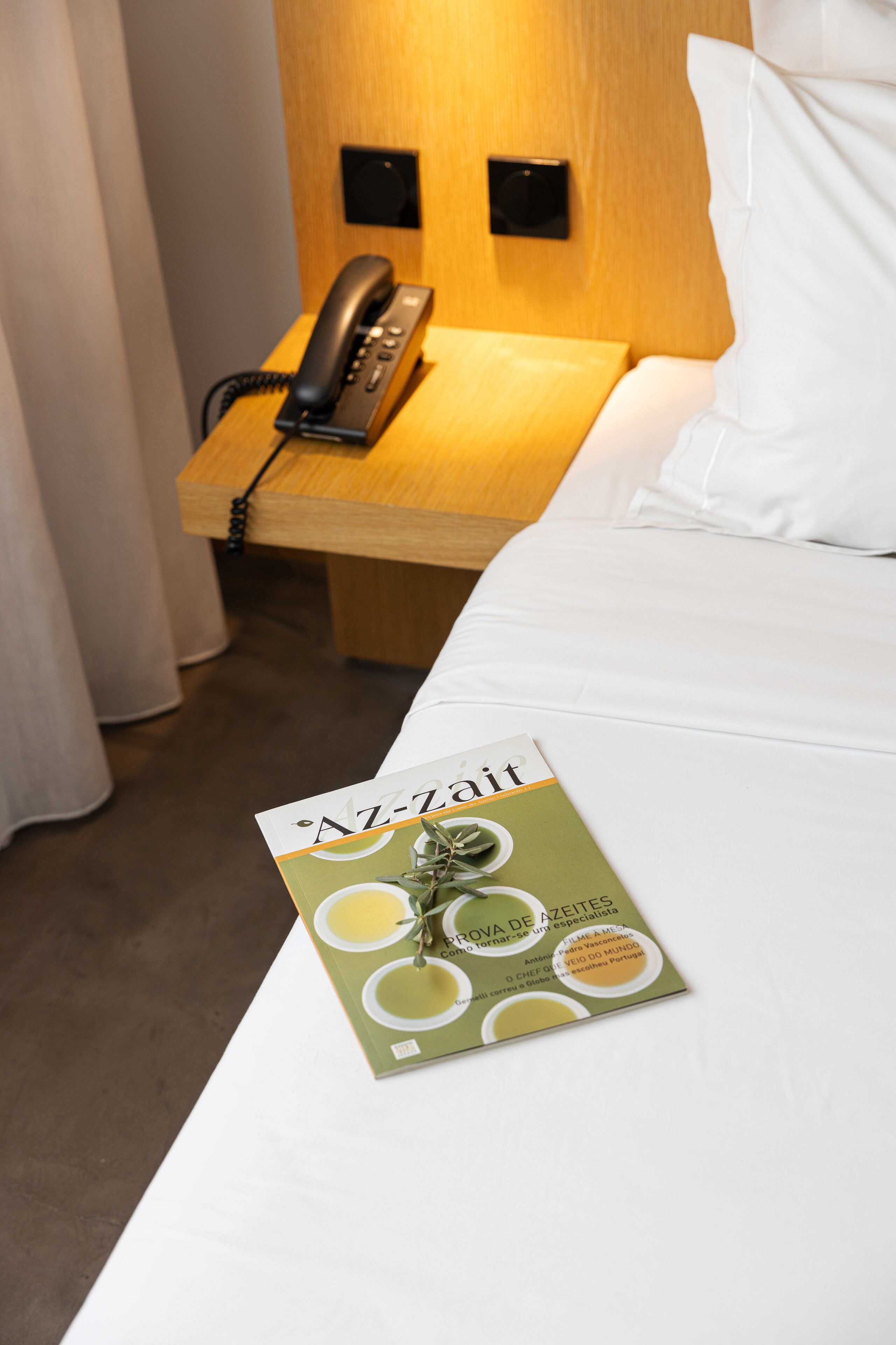 A hotel room with a wooden bedside table, a black telephone, and a brochure titled "Az-Zait".