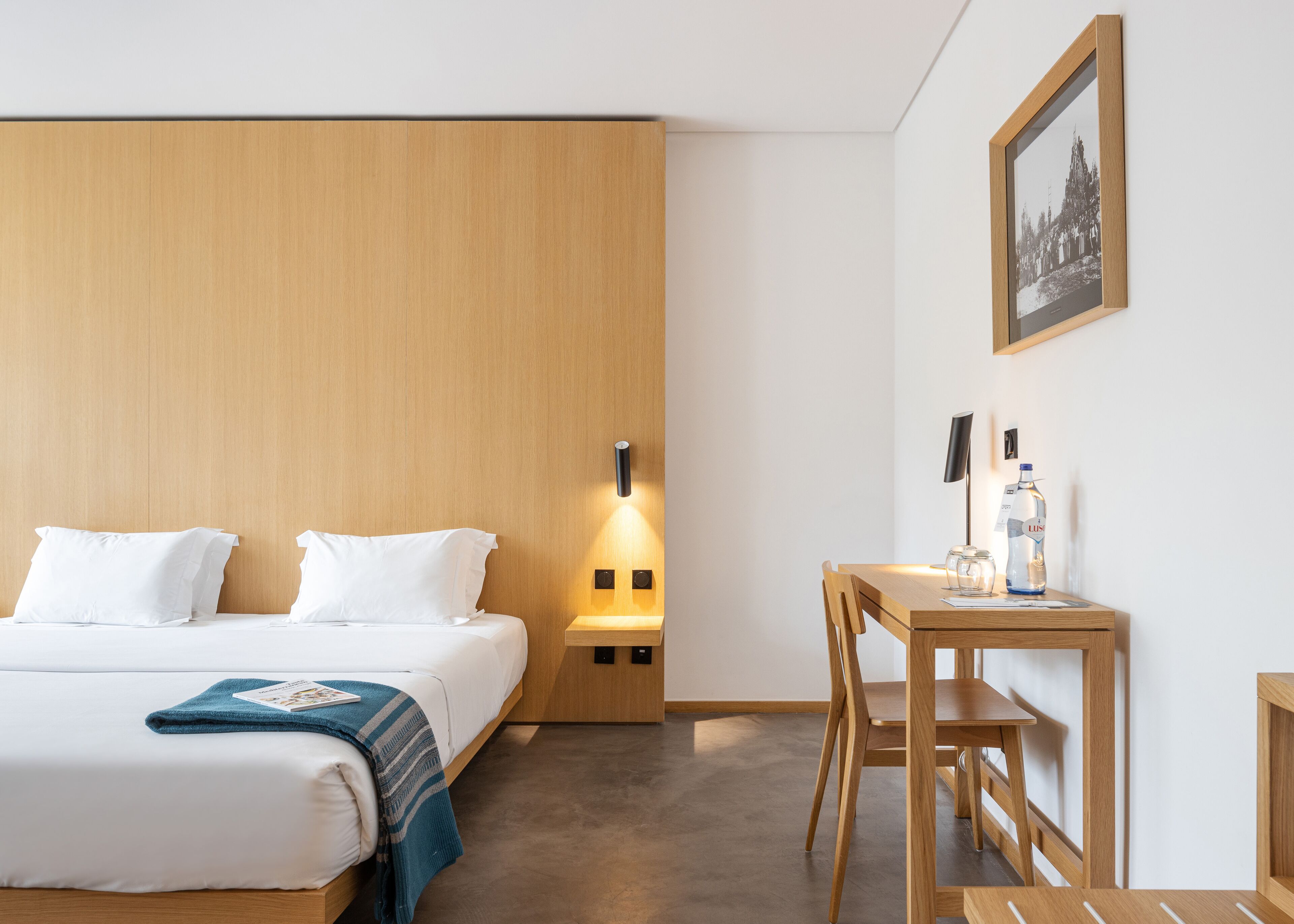 A hotel room with a bed, bedside table, lamp, and a framed picture on the wall.