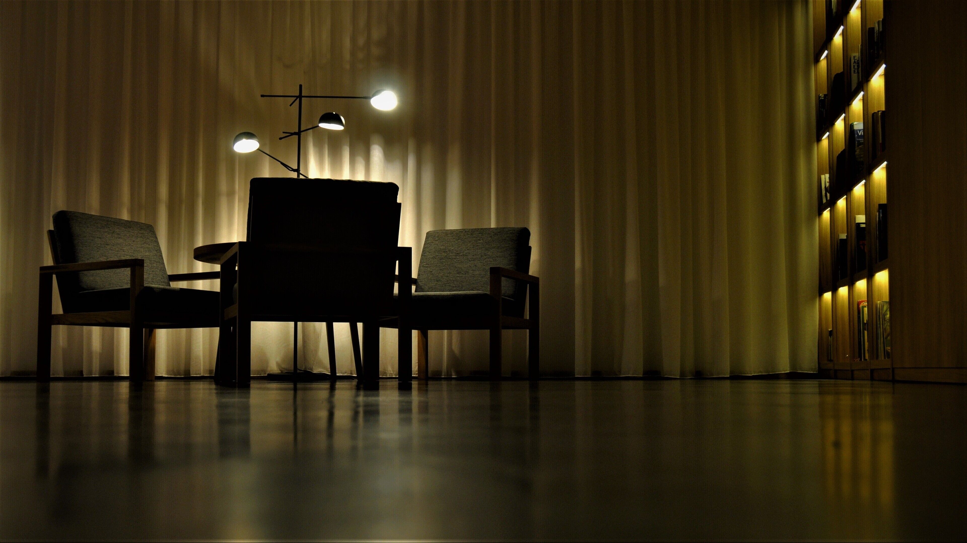 A dimly lit room with a table and two chairs, a curtain backdrop, and a bookshelf with illuminated shelves.