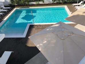 A swimming pool with a wooden deck, surrounded by white lounge chairs and umbrellas.