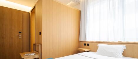 A hotel room with a bed, a wooden wardrobe, a small table, and a window with sheer curtains.
